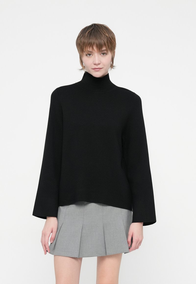 Musta poolpikkade varrukatega turtleneck-sviiter, millel on lõdva lõikega ja mis on valmistatud pehmest materjalist, koos halli volditud seelikuga.