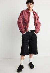 Νεαρός άνδρας με κόκκινο μπουφάν bomber πάνω από λευκό t-shirt με γραφικά, μαύρα σορτς μέχρι τα γόνατα, λευκές κάλτσες και μαύρα παπούτσια χωρίς κορδόνια, όρθιος με τα χέρια στις τσέπες.