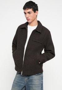 Braune Reißverschluss-Jacke mit Kragen, Fronttaschen und strukturiertem Stoff. Kombiniert mit einem weißen T-Shirt und Jeans.