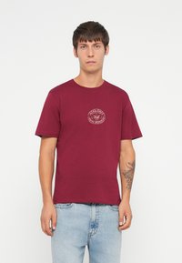 Burgunderfarget bomullst-skjorte med et sirkulært logo på brystet, korte ermer og rund hals, kombinert med lyseblå jeans.