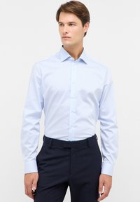 Uomo che indossa una camicia a righe blu chiaro e bianca con bottoni e pantaloni eleganti blu navy, in piedi contro uno sfondo chiaro uniforme, mentre guarda di lato.