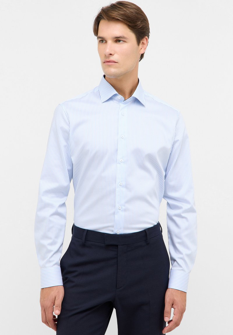 Uomo che indossa una camicia a righe blu chiaro e bianca con bottoni e pantaloni eleganti blu navy, in piedi contro uno sfondo chiaro uniforme, mentre guarda di lato.