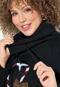 Sudadera negra con cuello alto tipo cowl y cordones. Presenta un diseño brillante de un corazón y un bastón de caramelo. Tejido suave y mangas ajustadas.
