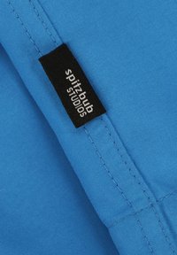 Blauwe stof met een gladde textuur, voorzien van een zwart label met witte tekst "spitz-bub STUDIOS." Zichtbare stiksels langs de rand.