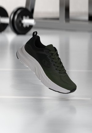 Scarpa da ginnastica verde scuro e nera con suola bianca spessa in palestra, manubrio sfocato sullo sfondo su pavimento riflettente.