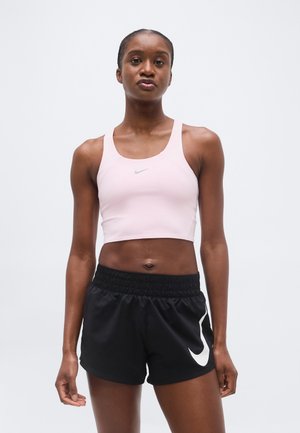 Fit vrouw staand, gekleed in een lichtroze Nike crop top en zwarte Nike sportshorts tegen een effen witte achtergrond.