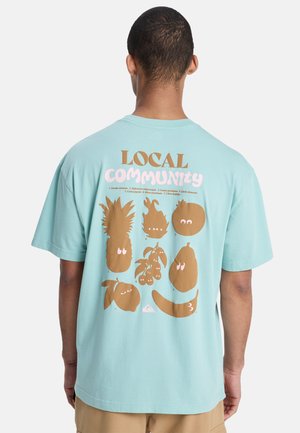 Osoba w jasnoniebieskiej koszulce z brązowymi owocami z kreskówkowymi oczami i napisem "LOCAL COMMUNITY" na plecach na jednolitym tle.