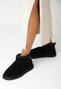 Černé semišové kotníkové boty s měkkou, kožešinovou podšívkou, kulatou špičkou a dekorativním prošíváním. Nosené s světle barevným pleteným šaty.