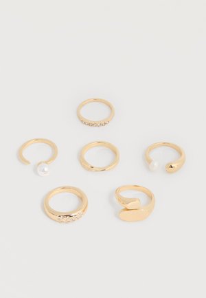 Seks guldringe med forskellige designs, inklusive åbne bånd med perler og bånd prydet med små krystaller, arrangeret på en hvid overflade.
