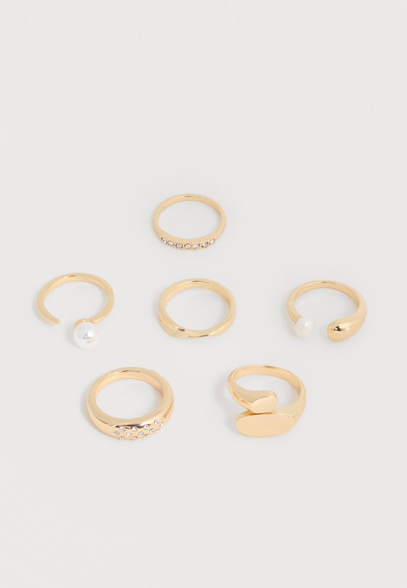 Έξι χρυσά δαχτυλίδια με διαφορετικά σχέδια, συμπεριλαμβανομένων ανοιχτών ζωνών με πέρλες και ζωνών διακοσμημένων με μικρούς κρυστάλλους, τοποθετημένα σε λευκή επιφάνεια.
