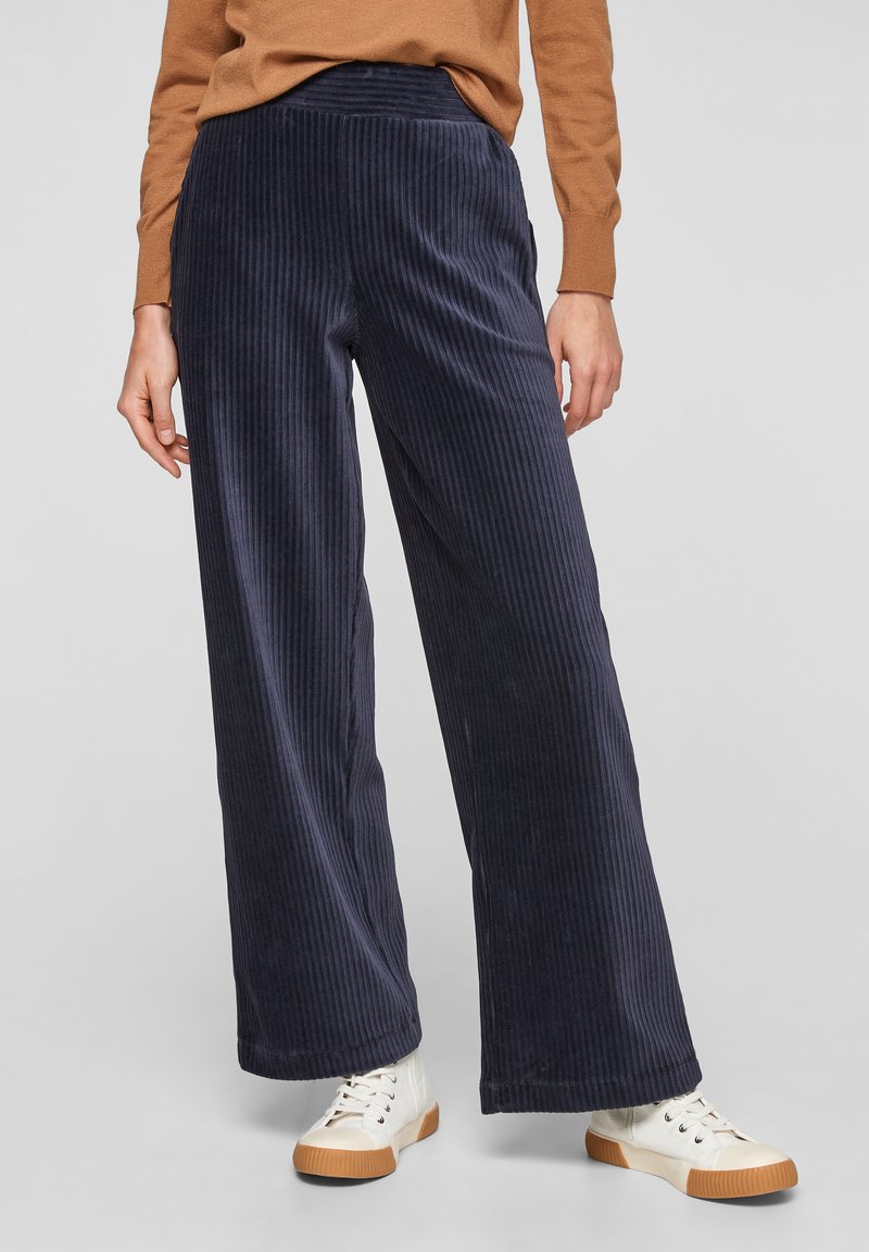 Femme portant un pantalon large en velours côtelé bleu marine, des baskets blanches et un haut marron à manches longues, debout devant un fond gris.