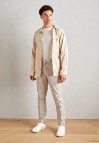 Giovane uomo che indossa una giacca beige aperta, una maglietta bianca, pantaloni con coulisse a righe beige e navy, e sneakers bianche, in piedi su un pavimento di legno.