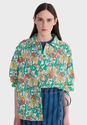 Mlada žena s ravnom smeđom kosom nosi šarenu cvjetnu košulju s gumbima preko tamnoplave majice i plave prugaste traper suknje.