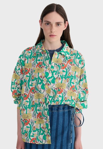 Jeune femme aux cheveux bruns raides portant une chemise à fleurs colorée boutonnée sur un haut marine et une jupe en denim rayée bleue.