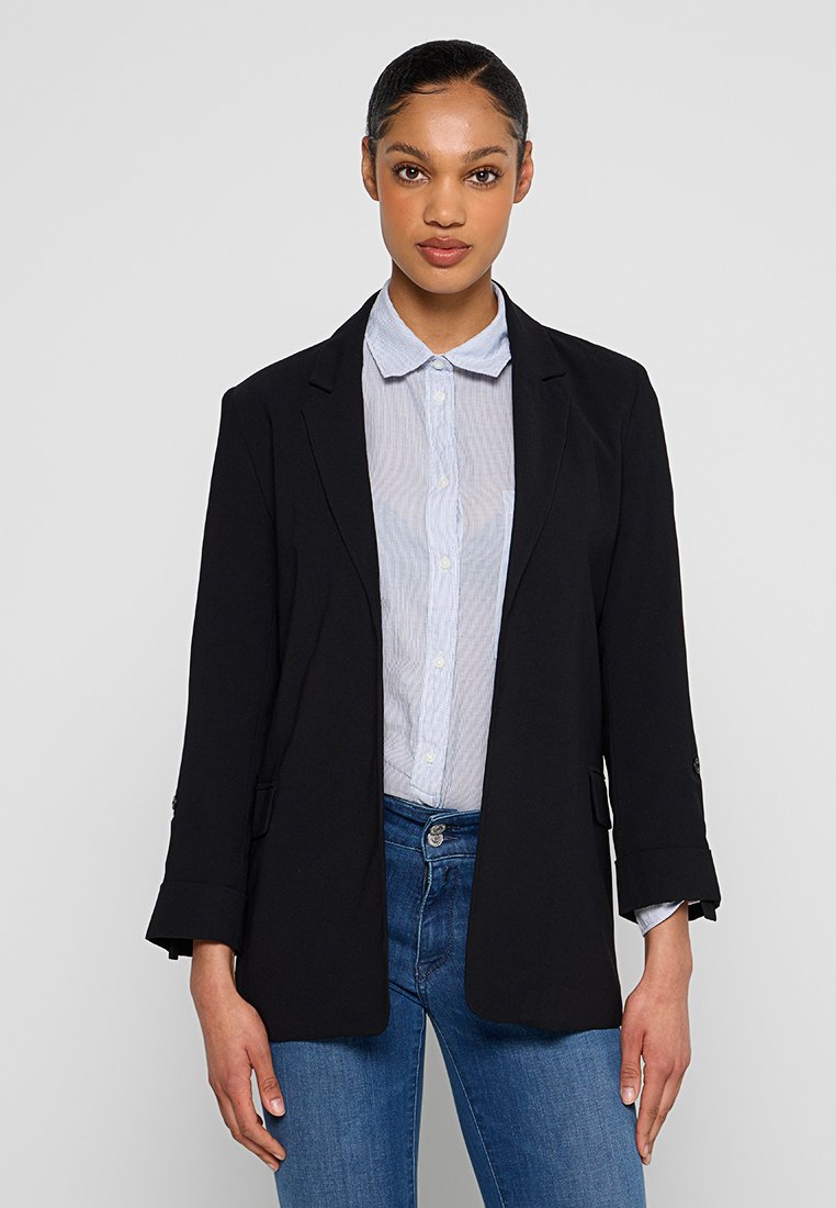 Blazer noir au design ajusté, tissu léger, revers crantés et manches retroussées, porté sur une chemise rayée bleu clair et un jean.