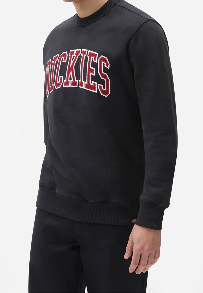 Černá mikina s žebrovaným výstřihem a manžetami. Na hrudi je výrazný červený vyšitý nápis "DICKIES". Hladká textura látky.