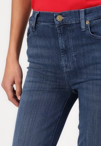 Blugi negri cu o croială slim, având un buton auriu și detalii de cusătură. Materialul pare a fi denim clasic cu o textură netedă.