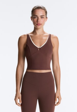 Jeune femme portant un crop top marron sans manches avec une bordure blanche et des leggings taille haute assortis, debout devant un fond uni.