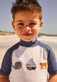 Weißes Kurzarm-Rashguard mit marineblauen Akzenten und bestickten Motiven eines Wals, Segelboots und einer Krabbe in Blau, Orange und Grau.