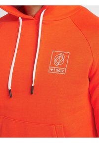 Sudadera naranja con cordones y acentos en los puños de color blanco. Presenta un logo gráfico en blanco en la zona del pecho. Tejido texturizado, corte relajado.