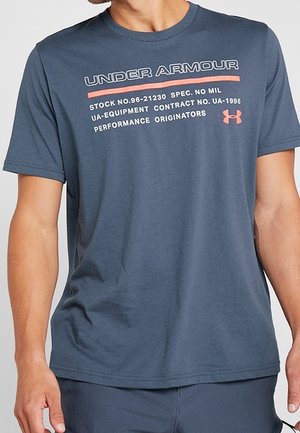 Homme portant un t-shirt bleu Under Armour avec un texte blanc et orange et un logo sur la poitrine, debout, les bras détendus le long du corps.