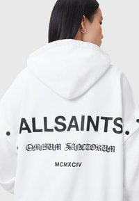 Sudadera blanca con texto negro en la parte posterior, con "ALLSAINTS" y otros detalles impresos. Incluye capucha y sutiles botones de acento en las mangas.