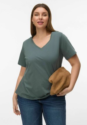 Woman wearing a green V-neck T-shirt and jeans, holding a soft brown clutch bag against a plain white background.