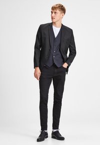Black suit jacket over a gray vest, paired with a white t-shirt and black pants. Wearing black shoes and white socks. Flat and tailored design.