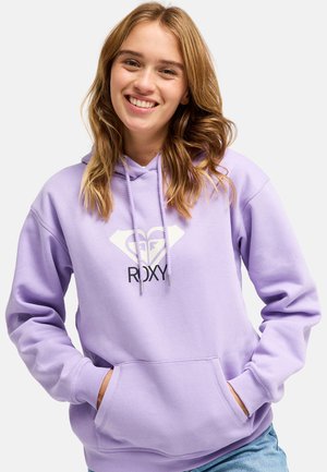 Młoda kobieta z jasnobrązowymi włosami uśmiechająca się, nosząca liliową bluzę z kapturem z białym i czarnym logo Roxy oraz rękami w przedniej kieszeni.