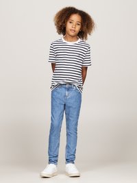 Gestreepte zwart-witte katoenen t-shirt, blauwe denim jeans en witte sneakers. Model staat met ontspannen armen en haar in losse krullen.