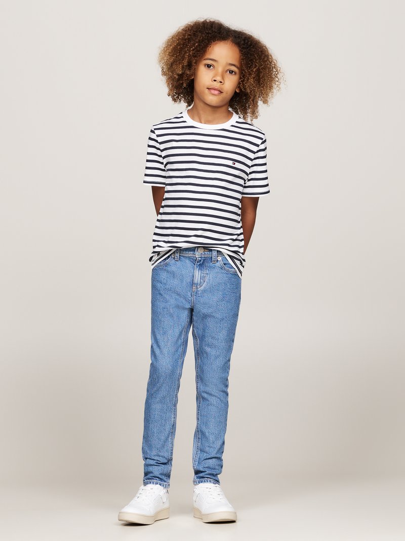 Gestreepte zwart-witte katoenen t-shirt, blauwe denim jeans en witte sneakers. Model staat met ontspannen armen en haar in losse krullen.