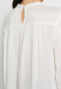 Blusa bianca e testurizzata con un tessuto leggermente trasparente, caratterizzata da piccole macchie in rilievo, pieghe e una chiusura a oblò sulla schiena con un bottone.