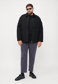 Zwarte canvas jas met een front-rits, gedragen over een zwart shirt. Grijze broek en zwarte sneakers maken de outfit af. Ronde zonnebril op.