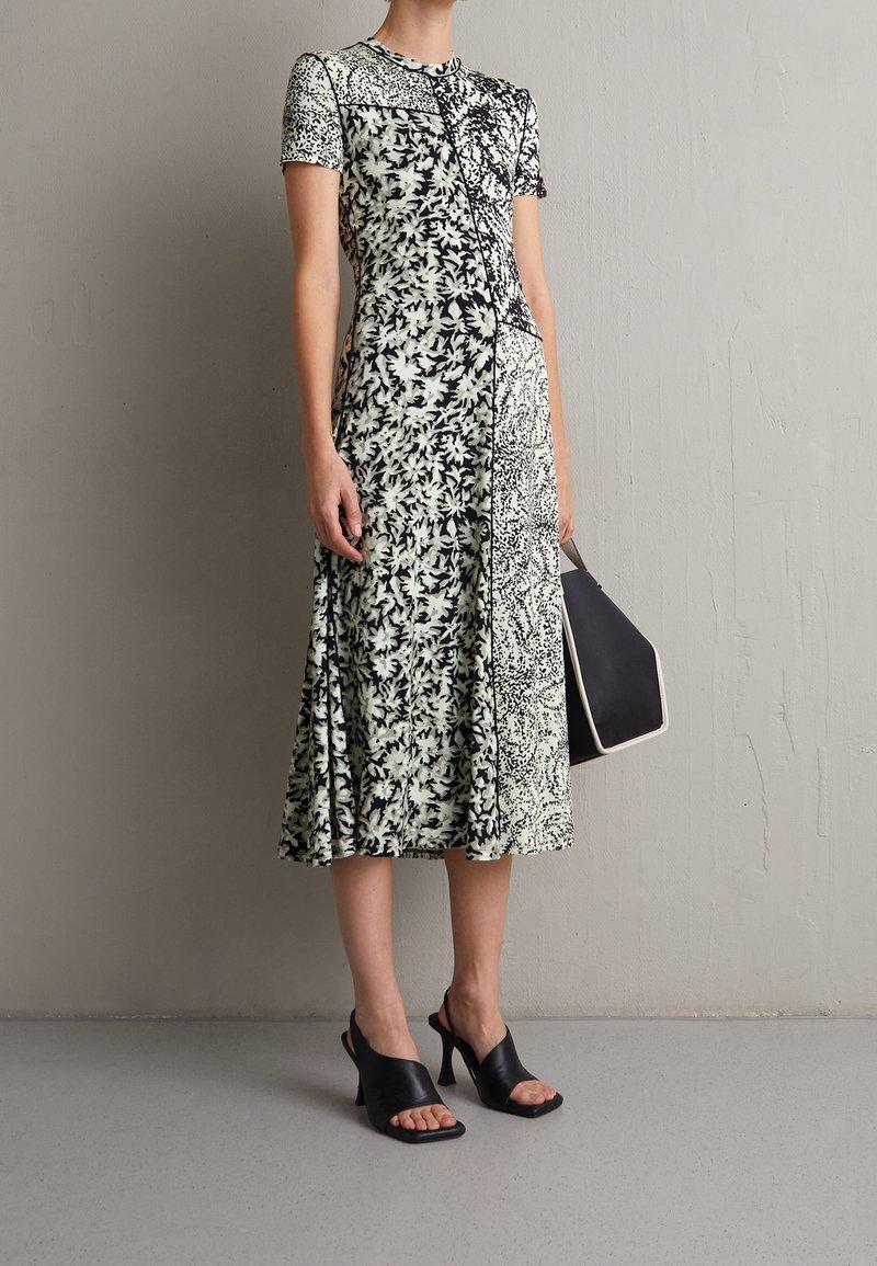 Sukienka midi z kwiatowym wzorem w kolorze czarnym i kremowym, krótki rękaw, dopasowany gorset, rozkloszowany dół, zestawiona z czarnymi sandałami na odkrytej pięcie i torebką.