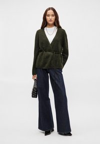 Zelený ovíječ s měkké látky, s páskem na uvázání a dlouhými rukávy, zkombinovaný s širokými džíny z tmavého denimu a kabelkou se cvočky.