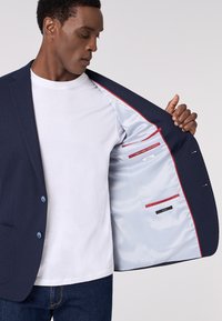 Blazer navy con tessuto testurizzato, design a una linea con bottoni blu. L'interno presenta una fodera chiara con accenti rossi e tasche.