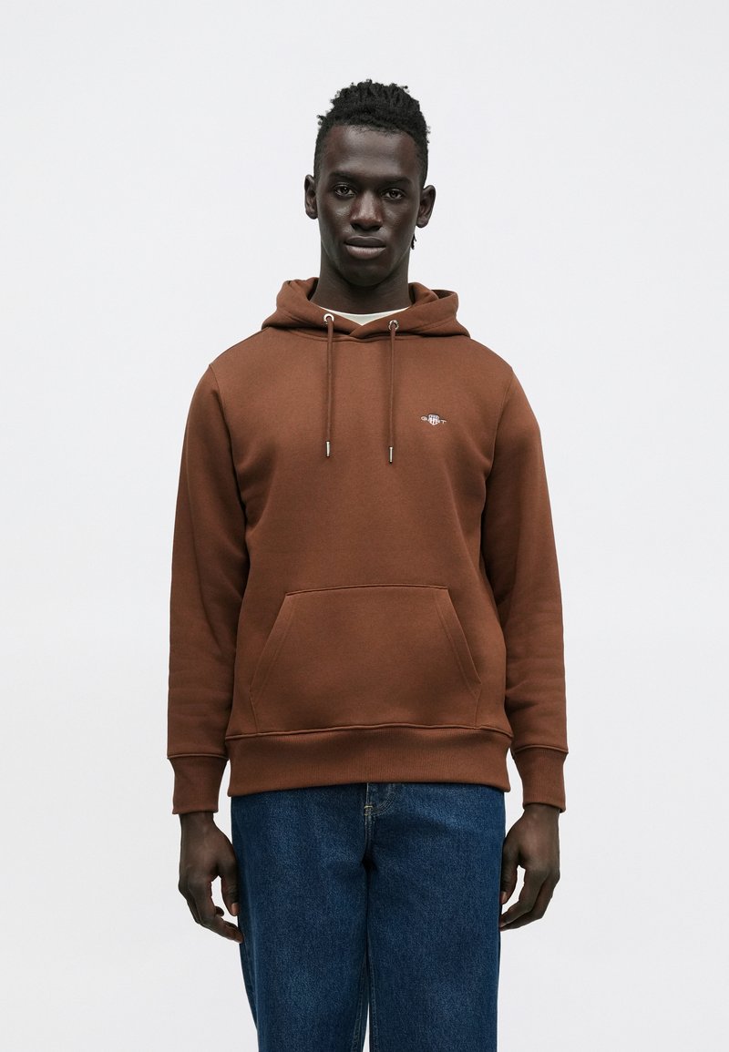 Barna kapucnis pulóver elülső zsebbel, húzózsinórokkal és egy kis logóval a mellrészen. Kék farmerrel párosítva, amely laza szabású.