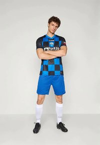 Jeune athlète masculin se tient les bras croisés, portant un maillot de soccer à carreaux bleu et noir, un short bleu, des chaussettes blanches et des crampons noirs.