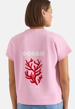Femme aux cheveux courts et bruns portant un t-shirt rose à manches courtes avec un corail rouge et le texte "ocean, in need of vitamin sea" dans le dos.