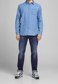 Camisa de mezclilla azul clara con mangas largas, cuello abotonado, un bolsillo en el pecho; combinada con jeans azul oscuro y zapatillas grises/verde.