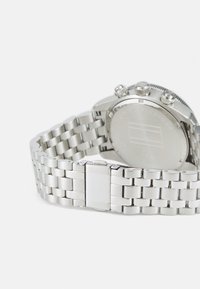 Montre en acier inoxydable argenté avec un bracelet en maillons métalliques, un boîtier rond et trois boutons chronographes. Logo gravé à l'arrière.