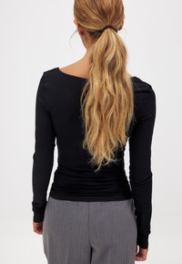 Top nero a maniche lunghe con scollatura rotonda, design aderente e texture liscia, abbinato a pantaloni plissettati grigi.