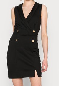 Femme portant une robe noire sans manches avec un décolleté en V profond, quatre boutons dorés et une petite fente sur le côté gauche.