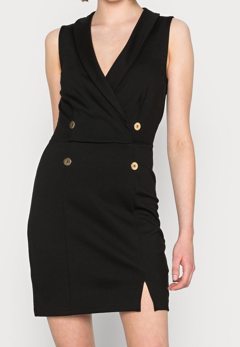 Femme portant une robe noire sans manches avec un décolleté en V profond, quatre boutons dorés et une petite fente sur le côté gauche.