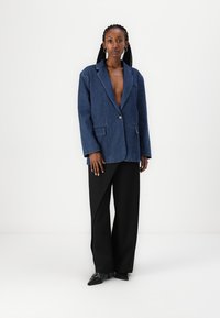 Denim blazer v temno modri barvi z enim gumbom za zapiranje, zarezanim ovratnikom in dvema sprednjima žepoma, v kombinaciji s črnimi širšimi hlačami.