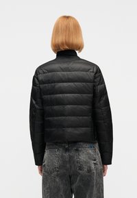 Άτομο με κοντά κόκκινα μαλλιά που φοράει μαύρο καπιτονέ μπουφάν και ξεθωριασμένο μαύρο τζιν, εμφανιζόμενο από πίσω σε απλό φόντο.