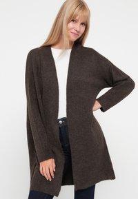 Cárdigan de punto marrón con un ajuste holgado, abertura frontal, mangas largas y textura suave. Llevado sobre una camiseta blanca y combinado con jeans oscuros.