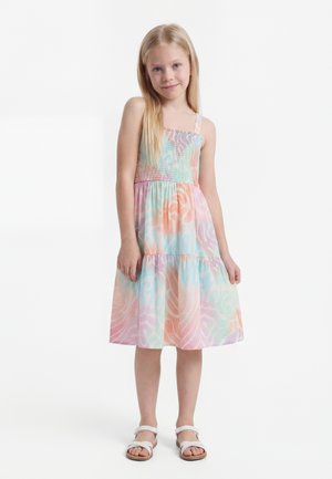 Nuori tyttö seisoo, päällä hihaton pastellinsävyinen kuviollinen mekko ja valkoiset sandaalit yksinkertaista valkoista taustaa vasten.