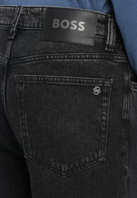 Črne kavbojke iz denima z usnjeno oznako BOSS na pasu, z zadnjim žepom z kovinskim logotipom in tradicionalnimi šivalnimi podrobnostmi.