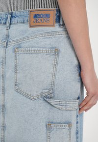 Svetlo modra denim krila z vidno šivom, zadnjimi žepi in rjavim etiketom z napisom "MOSCHINO JEANS" v modri barvi. Teksturna površina.